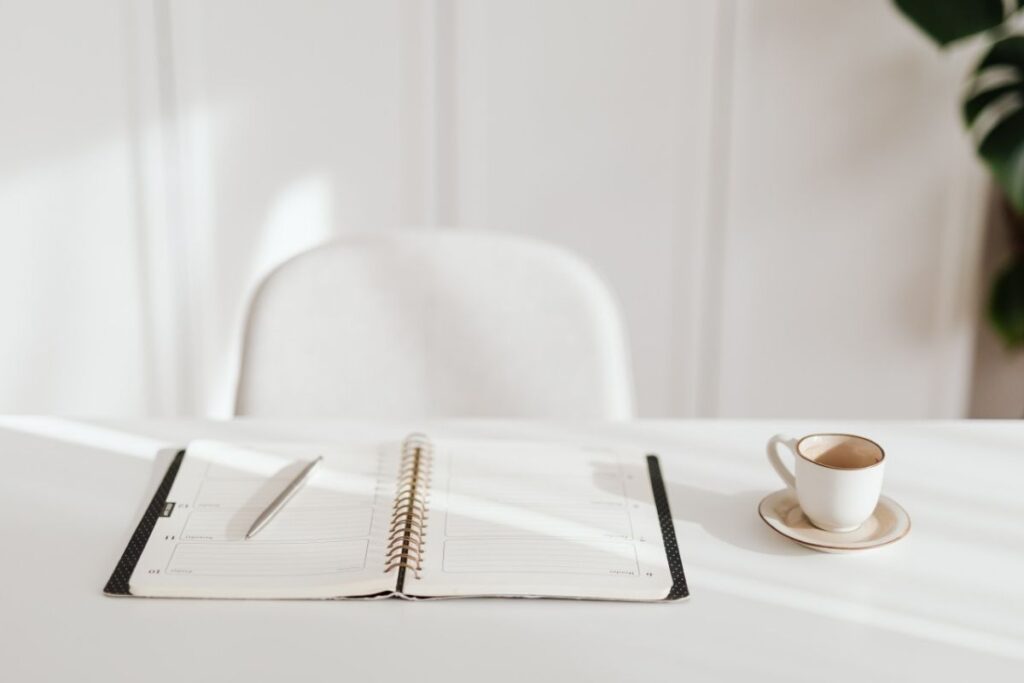 A white desk with a white chair and planner on the desk next to coffee.