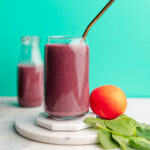 Banana orange celery smoothie sitting on two white marble coasters next to an orange and spinach leaves.